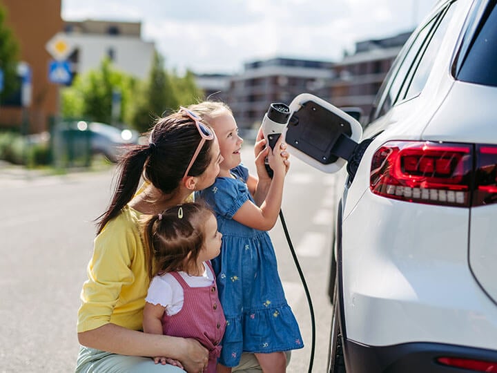 A mother with two young daughters helping one of them plug in an electric vehicle with a charger in a street charging port