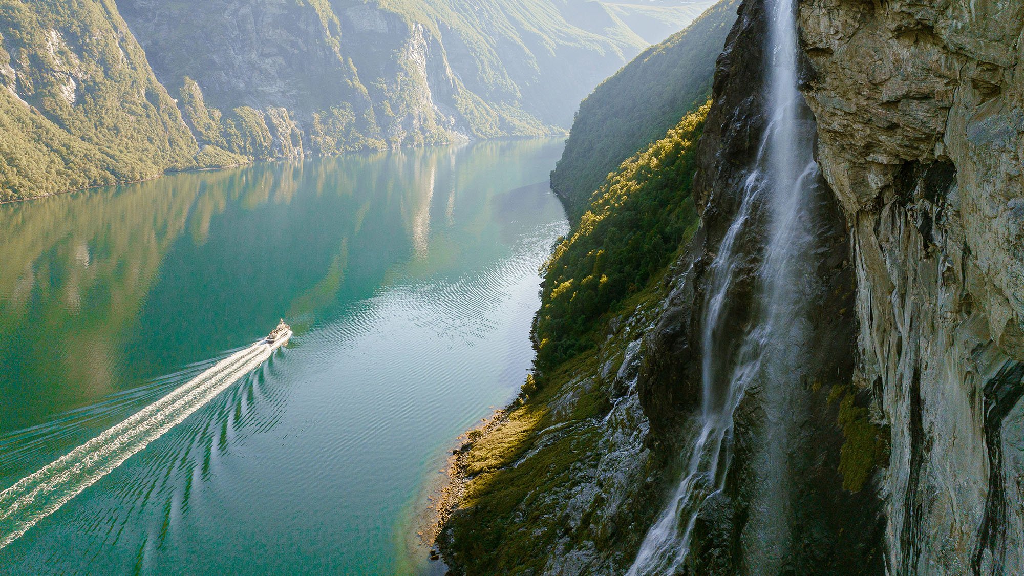 Ship on the water, passing through a canyon with a waterfall and mountains in view 